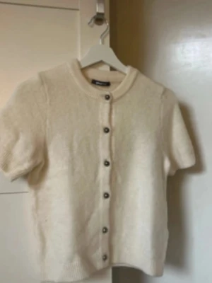 Stickad t-shirt Ginatricot  - Superfin cremefärgad kofta med korta ärmar och rund hals. Koftan har snygga knappar framtill och är stickad i ett mjukt material som känns lyxigt. Perfekt att slänga över en topp för en chill men ändå stilsäker look. Något nopprig, passar mer S