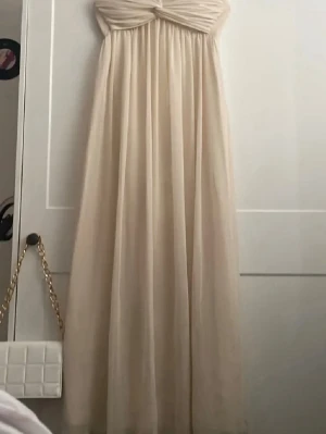 Champagnefärgad axelbandslös långklänning - Elegant axelbandslös långklänning i champagnefärg med twistad detalj upptill. Klänningen har ett mjukt, flytande fall och är tillverkad i ett tunt, skirt material som ger en lyxig känsla. Perfekt för speciella tillfällen där du vill känna dig extra fin.