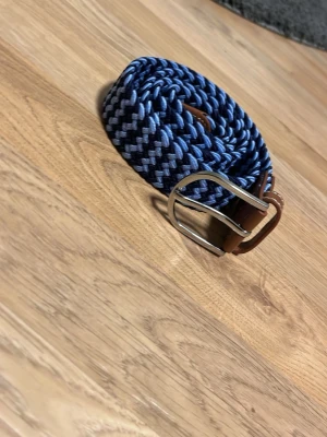 Blått flätat (Grish) bälte - Snyggt flätat bälte i marinblått och svart med bruna läderdetaljer och silverfärgat spänne. Perfekt accessoar för att lyfta din outfit med en cool och modern touch.