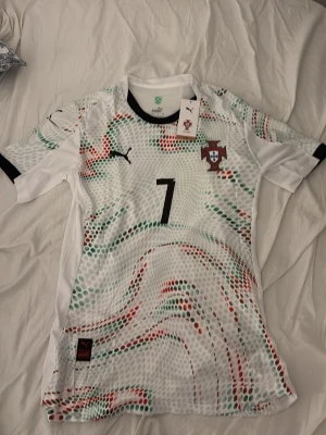 Portugal fotbollströja Puma #7 - Snygg Portugal fotbollströja från Puma med nummer 7. Tröjan är vit med gröna och röda mönster, svart Puma-logga och Portugals landslagsemblem på bröstet. Tillverkad i lätt och ventilerande material, perfekt för träning eller något snyggt till vardagen 