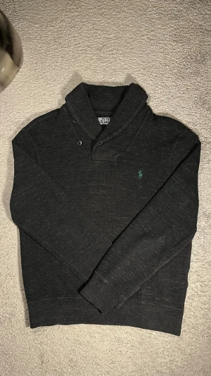 Mörkgrå stickad tröja Polo Ralph Lauren - Säljer en mörkgrå stickad tröja från Polo Ralph Lauren med klassisk logga broderad i grönt på bröstet. Tröjan har en snygg sjalkrage med knappar och långa ärmar. Perfekt för dig som vill ha en stilren och tidlös look.