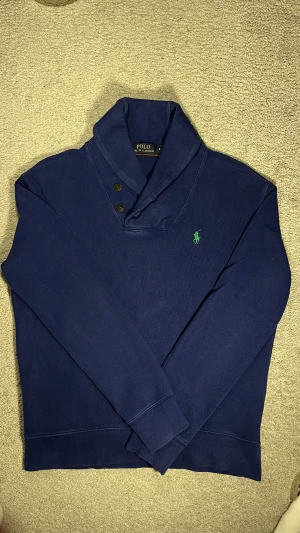 Marinblå stickad tröja Polo Ralph Lauren - Snygg marinblå stickad tröja från Polo Ralph Lauren med klassisk grön logga broderad på bröstet. Tröjan har en unik krage med två knappar och långa ärmar. Perfekt för dig som vill ha en stilren och tidlös look.