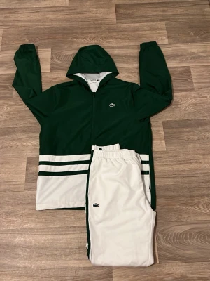 Grön och vit tracksuit från Lacoste - Snygg tracksuit från Lacoste i grönt och vitt. Jackan har huva, dragkedja och två vita ränder nertill. Byxorna är vita med gröna detaljer och har resår i midjan. Perfekt för träning eller chill. Klassisk Lacoste-logga på både jacka och byxor.
