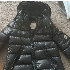 Svart dunjacka från Moncler - Snygg svart dunväst från Moncler med glansig finish och hög krage. Västen har dragkedja framtill, två dragkedjefickor och ikonisk Moncler-logga på bröstet. Perfekt för lager-på-lager och streetstyle. Storlek 3.