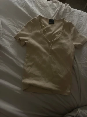 Beige ribbad topp från Ginatricot - Kortärmad beige topp från Ginatricot i ribbad bomull med v-ringning och knappar framtill. Perfekt basic-plagg med smickrande passform och mjukt material. Enkel att styla till olika looks.