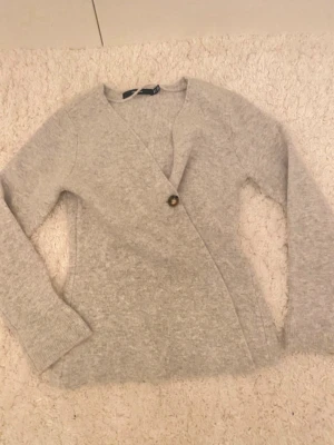 Ljusgrå omlottkofta med knapp - Mysig ljusgrå stickad omlottkofta med en dekorativ knapp framtill. Koftan har lång ärm och mjukt material som känns skönt mot huden. Perfekt för lager på lager och enkel att matcha med olika outfits.