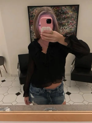 Svart transparent blus med volanger - Säljer en svart, transparent blus med långa ärmar och volangdetaljer längs halsringningen. Blusen har en lätt croppad passform och är gjord i ett tunt, luftigt material som ger en snygg och trendig look.