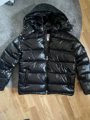 Svart glansig dunjacka från Moncler - Snygg svart dunjacka från Moncler med glansig finish och stor huva. Jackan har två dragkedjeförsedda fickor framtill och breda, vadderade paneler. Perfekt för kalla vinterdagar och riktigt trendig look. NFC tagg fungerar 