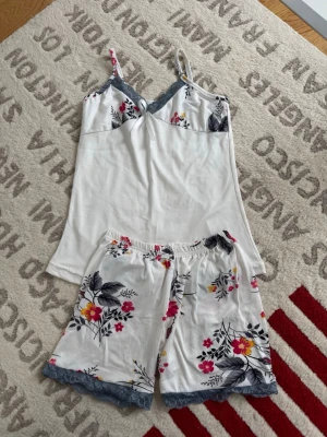 Blommig pyjamas med spetsdetaljer - Söt pyjamas med linne och shorts i vitt tyg med färgglatt blommönster i rosa, gul och grå. Linnet har tunna axelband och spetskant i blått vid bysten, medan shortsen har elastisk midja och blå spets vid bensluten. Perfekt för mysiga kvällar hemma.