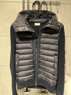 Mörkblå cardiganjacka från Moncler - Snygg mörkblå cardiganjacka från Moncler med vadderad front och huva. Ärmar och rygg är stickade i mjukt material medan framsidan har quiltad design. Tvåvägsdragkedja och fickor med tryckknappar. Perfekt för höst/vår men också sena sommarkvällar. Inga defekter så som hål eller missfärgningar, den är alltså i nyskick. Vid fler bilder eller frågor så är de bara att höra av dig! Pris kan diskuteras vid snabb affär. Ny pris ca 12 000.