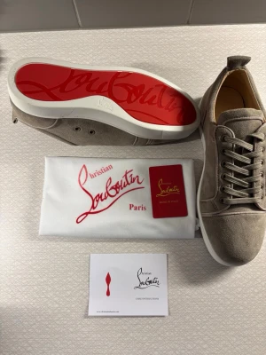 Beige mocka sneakers från Louboutin - Snygga beige sneakers från Christian Louboutin i mjuk mocka med klassisk röd sula och vita kanter. Skorna har rund tå, snörning och diskreta sömdetaljer. Perfekta för dig som vill ha en clean look med exklusiv känsla.