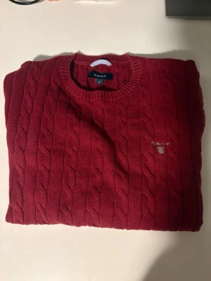 Röd kabelstickad tröja från GANT - Säljer en snygg röd kabelstickad tröja från GANT med rund hals och diskret logotyp broderad på bröstet. Tröjan har ribbade muddar och är tillverkad i mjukt stickat material som är perfekt för kyliga dagar. Säljer denna just för att den har blivit för liten. Hör av er vid minsta fundering😊