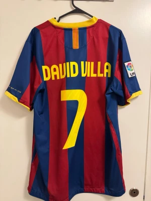 Barcelona David Villa matchtröja - Säljer en klassisk Barcelona matchtröja från Nike med David Villa och nummer 7 på ryggen. Tröjan har blå och röda vertikala ränder, gula detaljer och officiellt La Liga-märke på ärmen. Tillverkad i lätt och ventilerande material, perfekt för fotbollsträning eller samling., liten fläck på framsidan 