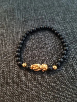 Svart pärlarmband med gulddetaljer - Snyggt armband med svarta pärlor och guldfärgade detaljer. Armbandet har en unik guldfärgad pärla i mitten och två mindre guldfärgade pärlor på varsin sida. Elastiskt band gör det enkelt att ta på och av. Perfekt accessoar för att lyfta din stil.
