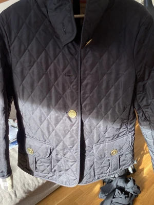 Svart quiltad rock från Burberry - Har ingen storlek men passar i allt från xs-mStilren svart quiltad rock från Burberry med klassiskt rutigt foder i beige, rött och svart. Rocken har guldfärgade knappar, två fickor med lock framtill och en hög krage. Perfekt för dig som vill ha en tidlös och snygg look.