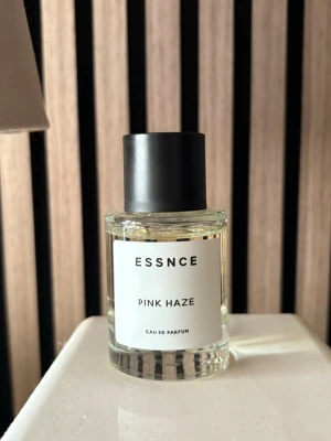 ESSNCE Pink Haze Eau de Parfum - Minimalistisk parfymflaska från ESSNCE med doften Pink Haze. Flaskan är cylindrisk i genomskinligt glas med svart matt kork och vit etikett med svart text. Innehållet är ljust gult och ger ett stilrent intryck, perfekt för dig som gillar enkel och modern design.