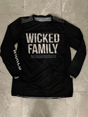Svart Crosströja Wicked Family - Säljer en svart långärmad sporttröja från Wicked Family med stora vita tryck fram och bak. Tröjan har texten 'WICKED FAMILY' på bröstet och nummer 788 samt namn på ryggen. Ventilerande meshpaneler på sidorna och detaljer på axlarna. Perfekt för motocross eller liknande sport. Nypris är 600kr.Det är i använt skick men inga synliga hål liknande 