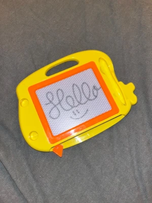 Ritplatta - Säljer en gul magnetisk ritplatta med orange ram och tillhörande penna. Perfekt för att rita eller skriva om och om igen utan papper. Plattan har ett handtag upptill och en suddfunktion med en orange spak längst ner- pennan är sladdfri. Inga defekter