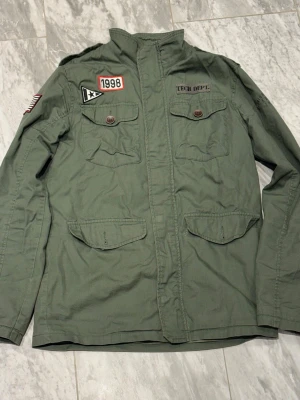 Grön vindjacka med patchar - Snygg militärgrön vindjacka med flera patchar, bland annat amerikansk flagga, '1998' och 'TECH DEPT.' på bröstet. Jackan har fyra fickor framtill med lock och knappar samt hög krage. Materialet är bomullstyg och jackan har en avslappnad passform.