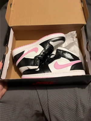 Nike Air Jordan 1 Mid rosa och svart - Snygga Nike Air Jordan 1 Mid sneakers i vitt, svart och rosa. Skorna har rosa swoosh och yttersula, klassisk vinge-logga på sidan och svartvita paneler i läder. Perfekta för dig som vill sticka ut med färg och ikonisk design.