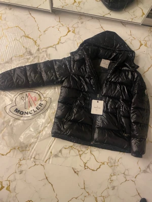 Svart dunjacka från Moncler med huva - Snygg svart dunjacka från Moncler med glansig finish och klassisk logotyp på ärmen. Jackan har två dragkedjefickor fram, huva och dragkedja hela vägen. Perfekt för kalla vinterdagar och riktigt trendig stil.