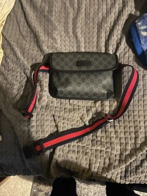 Gucci svart axelväska med monogram - Snygg svart axelväska från Gucci med klassiskt GG-monogram över hela väskan. Väskan har ett lock med magnetknäppning och justerbar axelrem i rött och marinblått. Tillverkad i canvas med läderdetaljer. Perfekt för dig som vill ha en stilren och trendig accessoar.