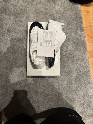 Maison Margiela svartvita sneakers - Unika sneakers från Maison Margiela i vitt läder med svarta detaljer och färgstänk på tå och sula. Klassisk låg modell med perforerade sidor och vita skosnören. Perfekt för dig som gillar statement-plagg och exklusiv design.