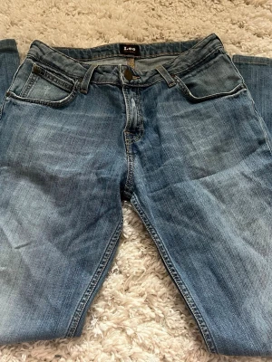 Blå raka jeans från Lee - Säljer ett par klassiska blå jeans från Lee med rak passform och fem fickor. Jeansen har normal midja, dragkedjegylf och snygga kontrastsömmar. Perfekta till en avslappnad stil och tillverkade i slitstark denim.
