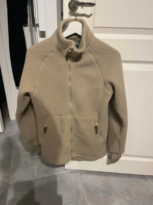 Beige teddyjacka med dragkedja - Mysig beige teddyjacka med hög krage och hel dragkedja framtill. Jackan har två fickor med dragkedja och muddar vid ärmsluten. Perfekt för kyliga dagar och ger en avslappnad, trendig look. Tillverkad i mjukt teddy-material.