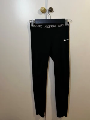 Svarta Nike Pro leggings - Säljer ett par svarta Nike Pro leggings med hög midja och elastisk linning där det står 'NIKE PRO'. Swoosh-logga på vänster lår. Tight passform och stretchigt material som passar perfekt till träning eller chill. Klassisk och sportig look.