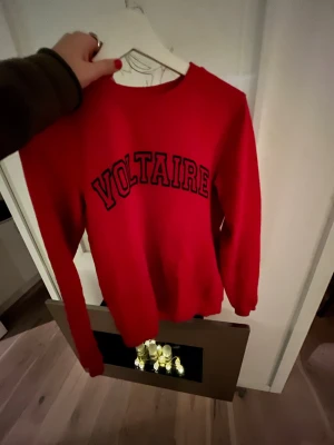 Röd sweatshirt från Voltaire - Snygg röd sweatshirt från Voltaire med svart text på bröstet. Tröjan har rund halsringning och långa ärmar. Perfekt för dig som gillar en enkel men trendig look. Materialet känns mjukt och bekvämt mot huden.