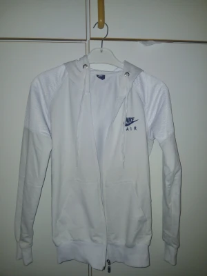 Vit Nike Air hoodie med dragkedja - Snygg vit hoodie från Nike Air med dragkedja framtill och huva med dragsko. Tröjan har ribbade muddar, två fickor och diskret Nike Air-logga i blått på bröstet. Tillverkad i mjukt material som känns skönt mot huden. Perfekt för en sportig och avslappnad stil.