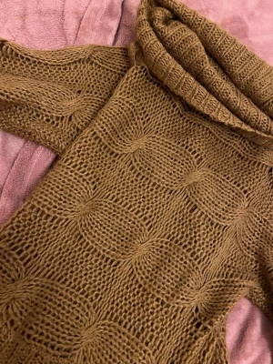 Brun stickad polotröja med mönster - Säljer en brun stickad polotröja med grovt kabelstickat mönster. Tröjan har lång ärm och en hög, ribbad polokrage. Materialet känns mjukt och mysigt, perfekt för kyliga dagar. Ingen synlig logga eller märke.