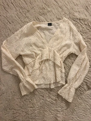 Vit strukturerad blus Gina Tricot - Säljer en vit blus från Gina Tricot med strukturmönster och volangdetalj framtill. Blusen har v-ringning och långa ärmar med resår vid ärmslut. Materialet är lätt och transparent, perfekt för en trendig look.