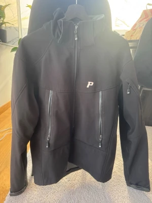 Svart vindjacka från Peak Performance - Snygg svart vindjacka från Peak Performance med dragkedja framtill, två vertikala bröstfickor med vattentäta dragkedjor och en liten logga på bröstet. Jackan har huva och är tillverkad i ett slätt, tekniskt material som står emot vind.