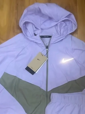 Ljuslila Nike hoodie med dragkedja - Snygg ljuslila vindjacka från Nike med dragkedja framtill och huva. Har en kontrasterande grön diagonal detalj över bröstet och den klassiska Nike-loggan i vitt på ena sidan. Perfekt för en sportig och avslappnad stil.