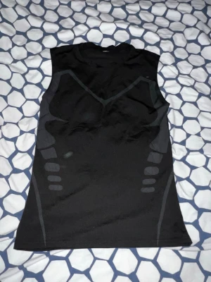 Svart ärmlös träningströja - Säljer en svart ärmlös träningströja med diskreta gråa mönster på både fram- och baksida. Tröjan är tillverkad i ett stretchigt funktionsmaterial som andas, perfekt för gymmet eller löpning.