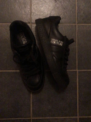 Svarta Versace Jeans Couture sneakers - Snygga svarta sneakers från Versace Jeans Couture med logga i vitt på sidan och tungan. Skorna har klassisk låg profil, svarta skosnören och är tillverkade i slätt läderliknande material. Perfekta för dig som vill ha en stilren och trendig look.