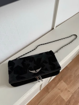 Svart axelväska från Zadig & Voltaire - Cool svart axelväska från Zadig & Voltaire med sammetsstjärnor och silverfärgad kedjerem. Väskan har ett snyggt vingemblem i metall framtill och dragkedja. Lite slitage på dragkedjan annars som ny!