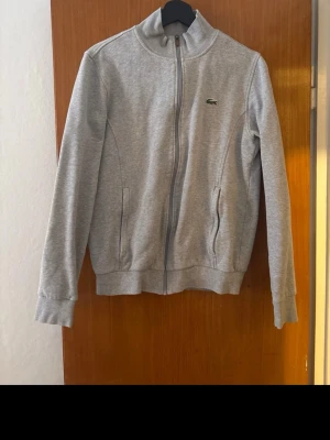 Grå zip hoodie från Lacoste - Snygg grå zip från Lacoste med dragkedja framtill och två fickor. Klassisk krokodil-logga på bröstet och ribbade muddar vid ärmar och nederkant. Tillverkad i mjukt sweatshirtmaterial som är riktigt skönt att ha på sig.