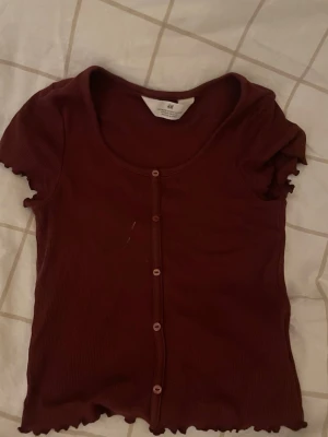 Vinröd ribbad topp från H&M - Snygg vinröd ribbad topp från H&M med korta ärmar och knappar framtill. Toppen har en croppad passform och vågig kant vid ärmslut och nederkant. Perfekt att styla med jeans eller kjol för en trendig look.