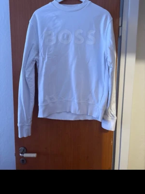 Vit sweatshirt från Hugo BOSS - Snygg vit sweatshirt från BOSS med diskret logga i relief på bröstet. Tröjan har rund halsringning, långa ärmar och ribbade muddar vid ärmslut och nederkant. Perfekt för en clean och stilren look.