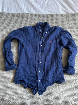 Blå linneskjorta från Ralph Lauren - Snygg blå linneskjorta från Ralph Lauren med vita knappar. Skjortan är långärmad och har den ikoniska röda loggan broderad på bröstet. 