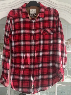 Röd rutig skjorta Legendary Whitetails - Klassisk röd och svart rutig skjorta från Legendary Whitetails, modell Buck Camp. Skjortan har långa ärmar, bröstficka med knapp och knäppning framtill. Tillverkad i mjuk bomull med insida i manchester för extra detalj.