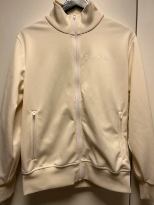Beige zip hoodie från Palm Angels - Snygg beige hoodie från Palm Angels med dragkedja framtill och två fickor med dragkedja. Diskret Palm Angels-logga broderad på bröstet och vita samt svarta ränder längs ärmarna. Tillverkad i mjukt material som känns skönt mot huden. Perfekt för en avslappnad streetwear-look.