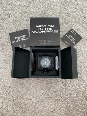 Omega x Swatch MoonSwatch Moonphase - Säljer en Omega x Swatch MoonSwatch Mission to the Moonphase Snoopy med svart biokeramiskt boett och svart textilband. Klockan har vit urtavla med detaljer i silver och flera funktioner som kronograf och tachymeter. Levereras i originalförpackning med tillbehör.