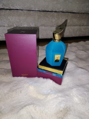 Xerjoff blå parfymflaska med guldlock 100ml - Lyxig parfym från Xerjoff i en unik blå flaska med sammetsliknande yta och elegant guldfärgat lock. Flaskan har en guldfärgad etikett framtill och kommer i en lila och guld presentförpackning. Perfekt för dig som gillar exklusiva dofter och snygg design.