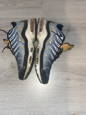 Nike Air Max Plus TN grå/guld - Nike Air Max Plus TN sneakers i grått och guld med svarta detaljer. Skorna har synliga Air Max-enheter i sulan, mesh- och syntetmaterial på ovandelen samt vita och svarta inslag. Ikonisk TN-logga på plösen och sidorna.