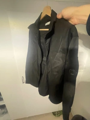 Svart overshirt från C.P. Company - Säljer en svart overshirt från C.P. Company med dragkedja framtill, bröstficka och den ikoniska linsen på ärmen. Som ni ser så är batchen tyvärr lite sliten då jag råkat skrapa emot, utöver de så är den i super fint skick.