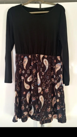 Svart paisleymönstrad klänning - Svart klänning med lång ärm och rund halsringning. Kjoldelen har ett paisleymönster i beige, brunt och vitt med inslag av blått. Överdelen är enfärgad och kjolen har en lätt utställd form. Perfekt för dig som gillar boho-vibbar.